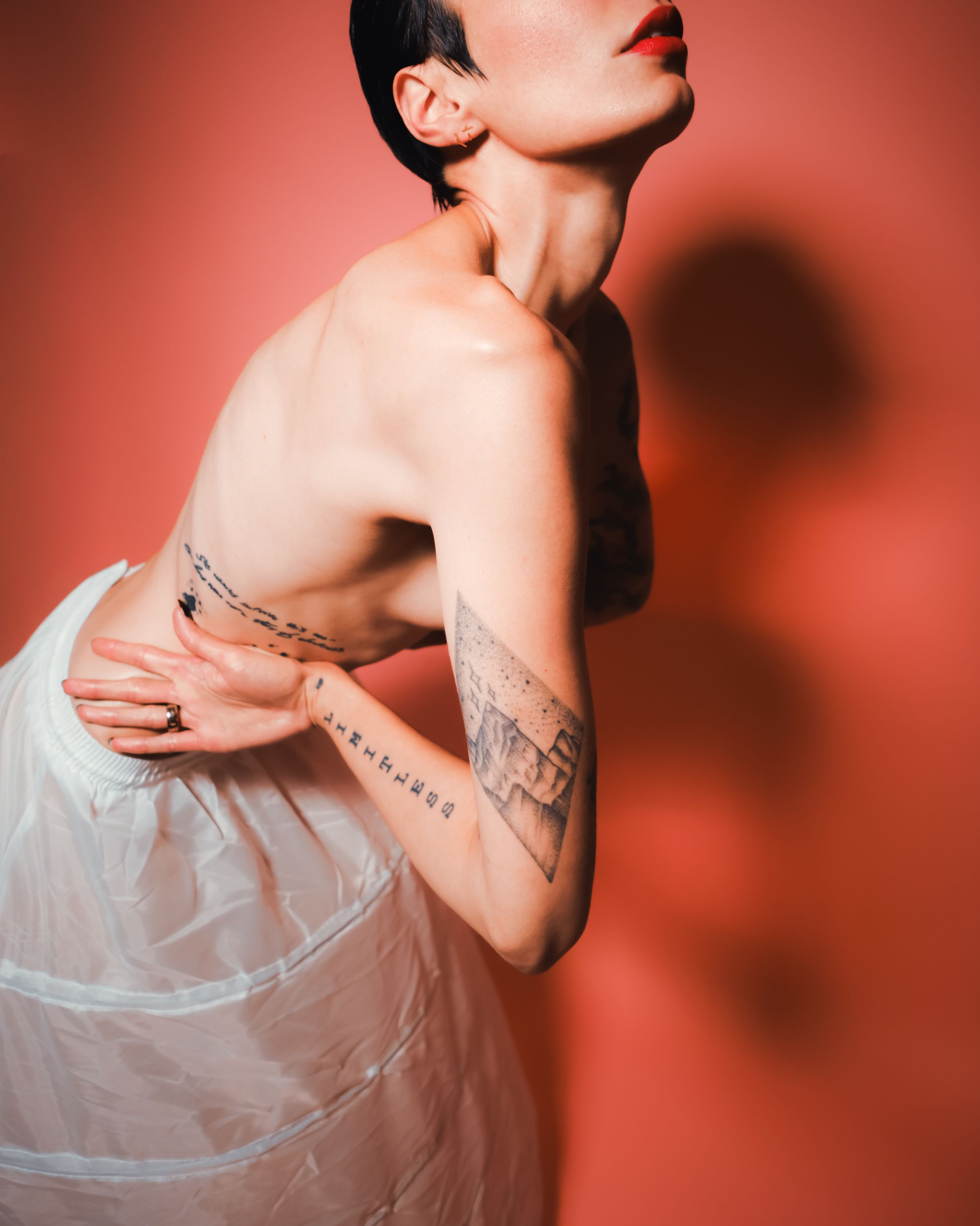 Editorial portrait of Alia Kohlmann leaning slightly forward with one arm across her chest, captured against a coral backdrop by Dario Hudon