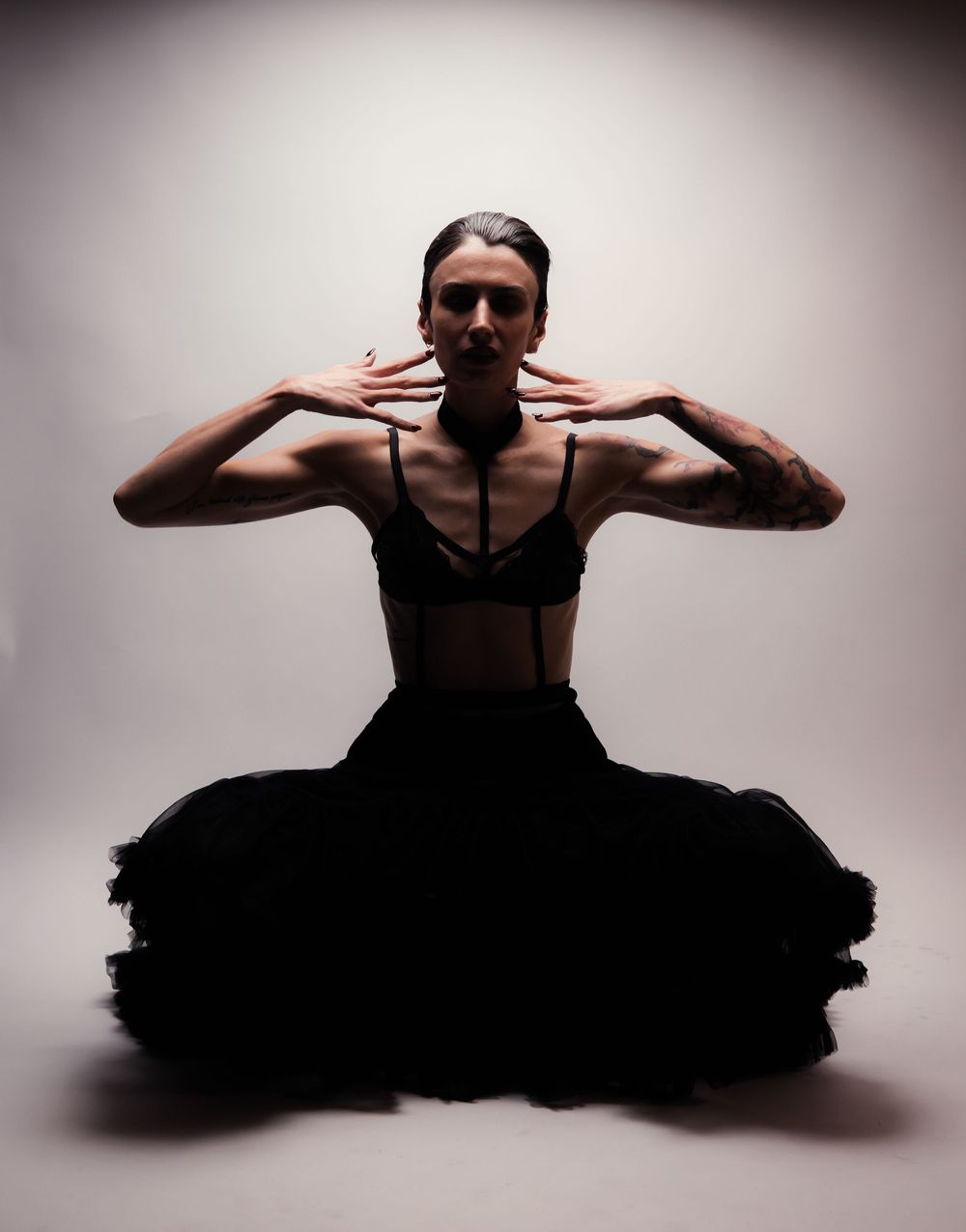Studio editorial portrait of Alia Kohlmann kneeling in a black skirt, raising her arms near her shoulders under dramatic lighting.